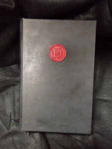 A smooth black leather book with black velvet bookmark and an ornate red wax seal on the cover, sitting on a crinkled black background.